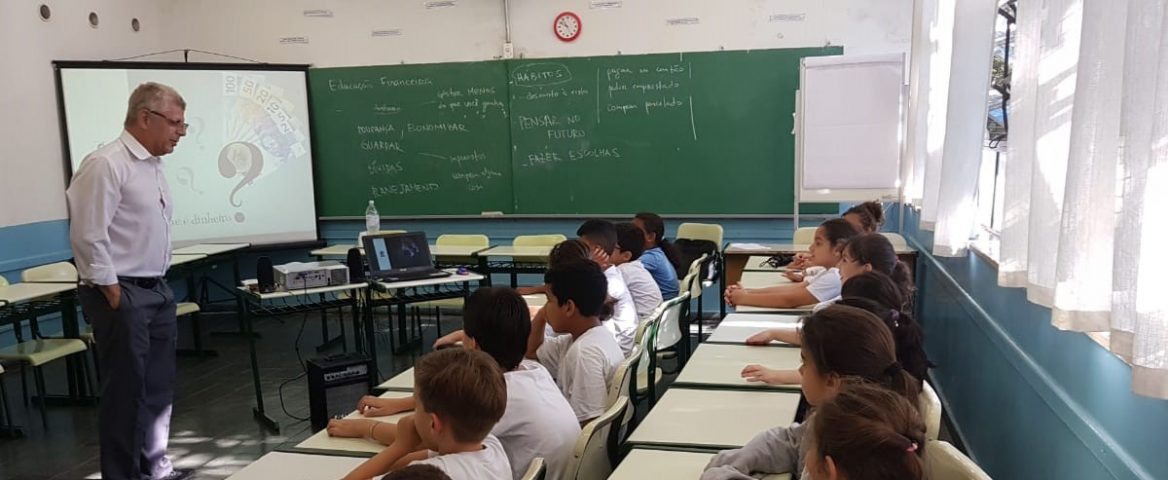 Crianças sentadas em carteiras, em uma sala de aula, olhando para o professor, que está ao lado de um telão. Ao fundo, há uma lousa.