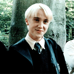 Draco Malfoy está em frente a uma árvore, numa cena externa. Ele remove a alça de sua bolsa de seu ombro, ao mesmo tempo que sorri de forma irônica. 