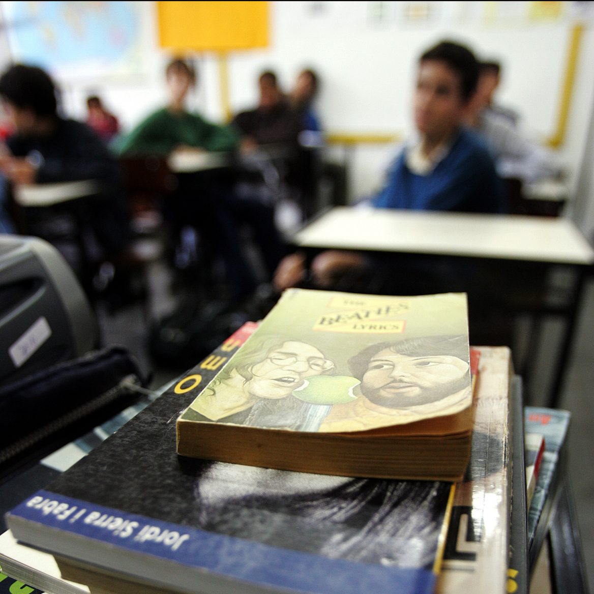 O foco da imagem são livros em cima de uma mesa. Ao fundo, estudantes sentados em carteiras de sala de aula.