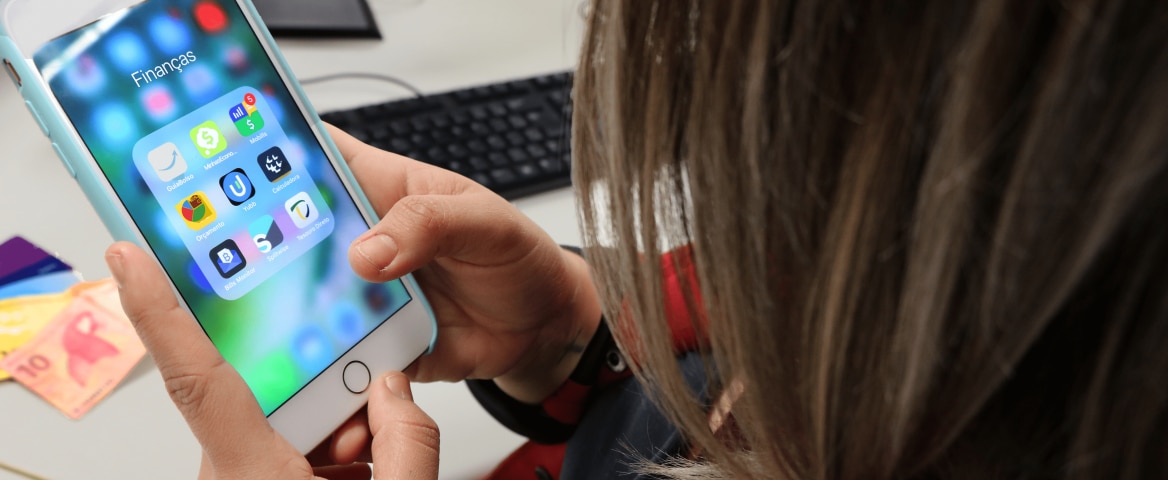 Jovem segura um celular, cuja tela está aberta em uma pasta de aplicativos sobre finanças. Ao fundo, estão cartões de crédito, dinheiro e a tela de um computador.