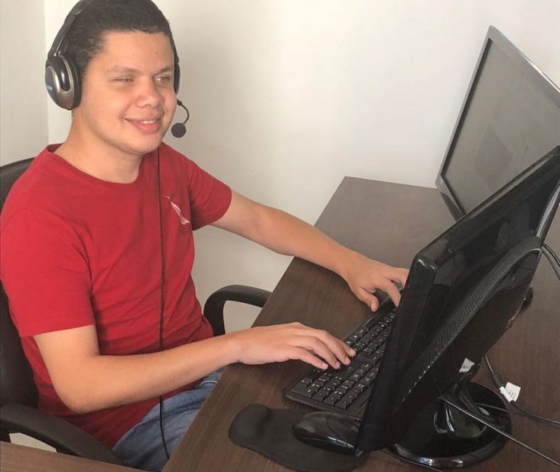 Wesley Gamaliel está sentado, usando um computador com duas telas. Ele veste camiseta vermelha, calça jeans e fones de ouvidos que cobrem as orelhas. Ele está com as mãos no teclado.