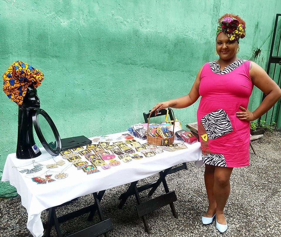 Michele Fernandes, mulher negra, posa para foto ao lado de peças de roupas e acessórios criados e confeccionados por ela mesma. Entre eles, turbantes, brincos e pulseiras.