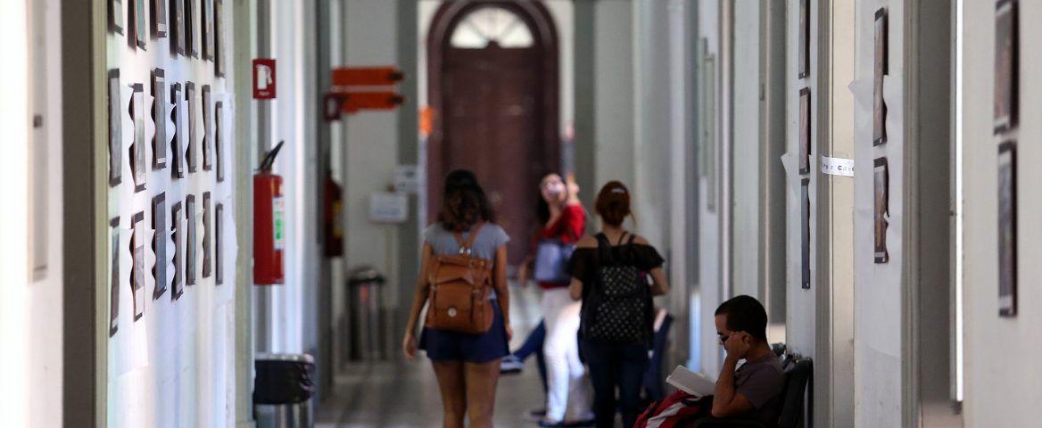 Corredor de um dos prédios da Escola de Comunicação da UFRJ. O corredor é alto, de um prédio antigo. Ao fundo, duas estudantes caminham, de costas para a lente da câmera. Um estudante, um pouco mais a frente, está sentado num banco de madeira lendo. Outra pessoas mais ao fundo, mas pouco visíveis.